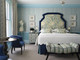 A vibrant blue and green bedroom suite at Covent Garden Hotel, featuring striped wallpaper, a botanical headboard, and a footstool upholstered in Romany Weave Aqua by Kit Kemp Design Studio.